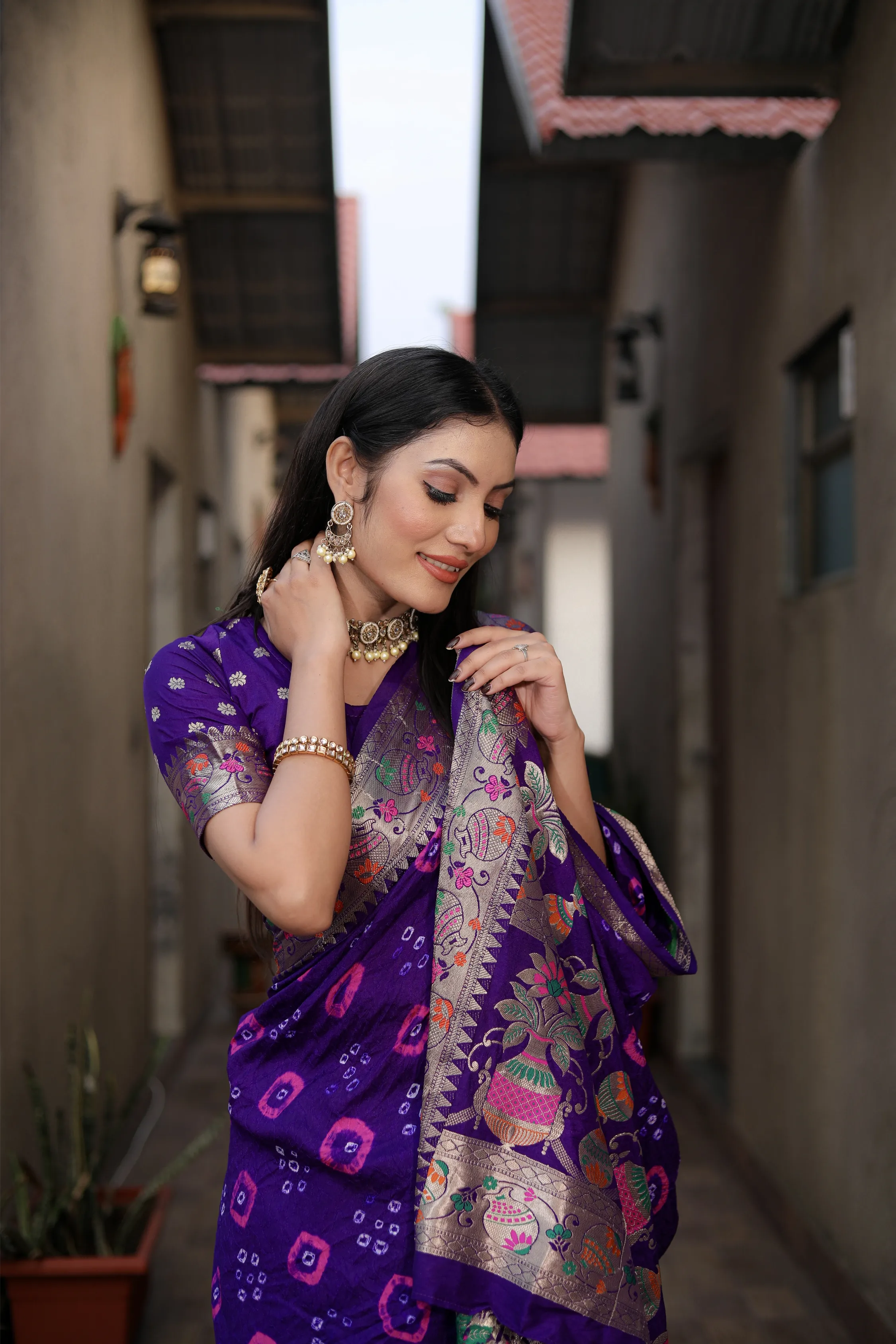 Woman in a purple saree with intricate patterns standing in an alleyway.