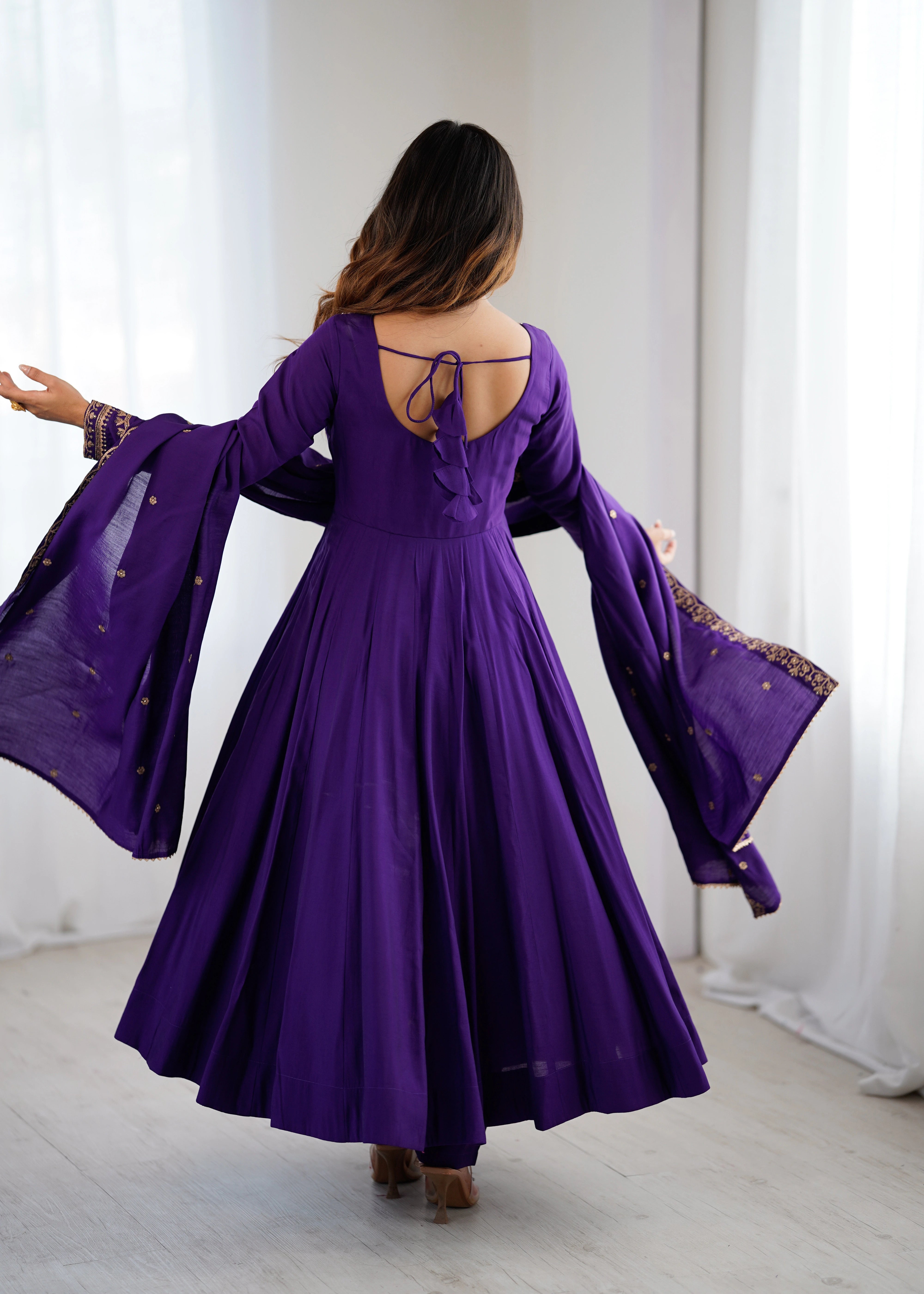 Woman wearing a purple dress with long sleeves and a backless design, standing in a room with white curtains.