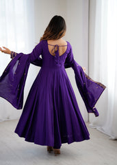 Woman wearing a purple dress with long sleeves and a backless design, standing in a room with white curtains.