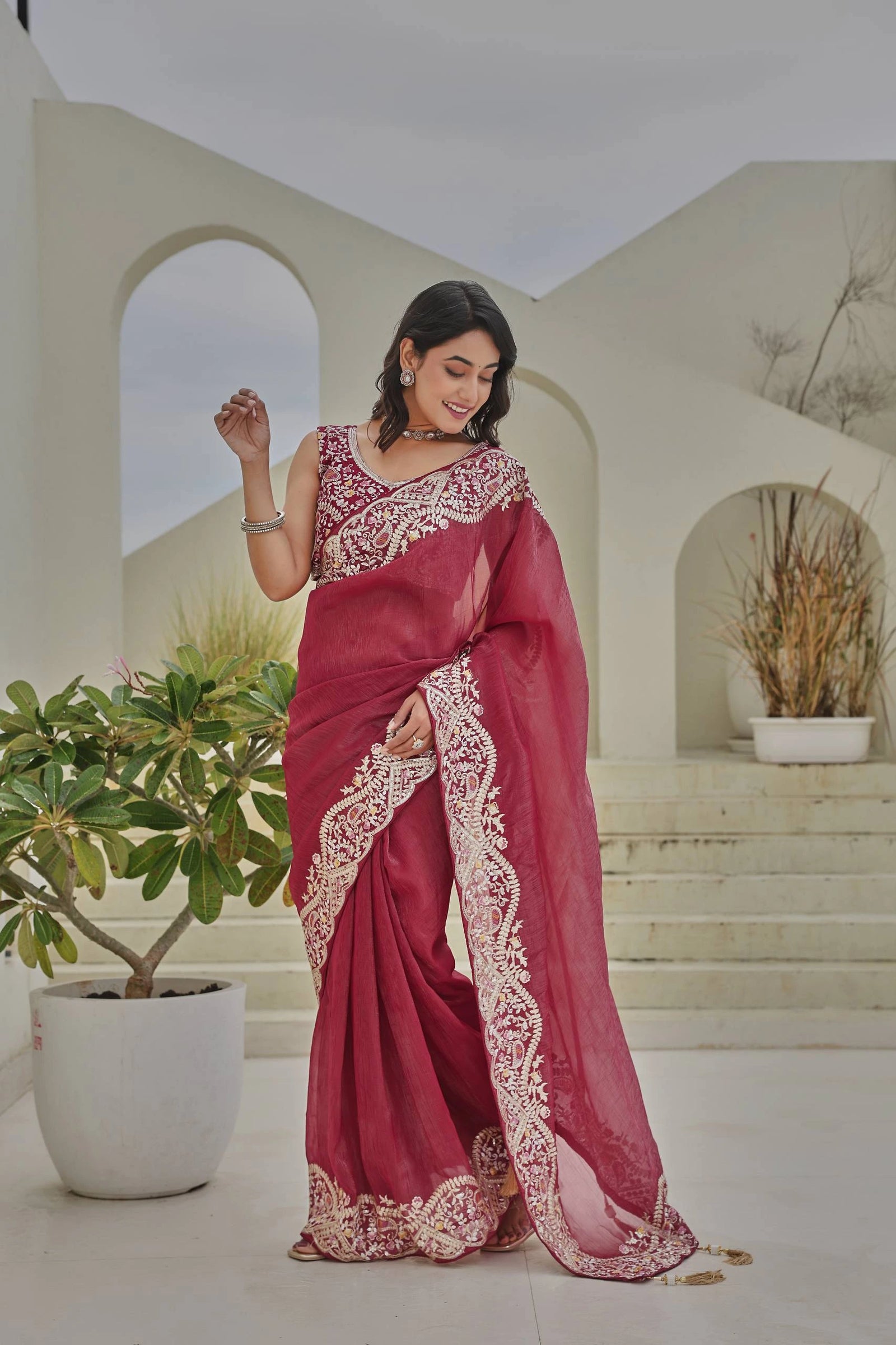 Woman in a red saree with white embroidery standing in an outdoor setting with plants.