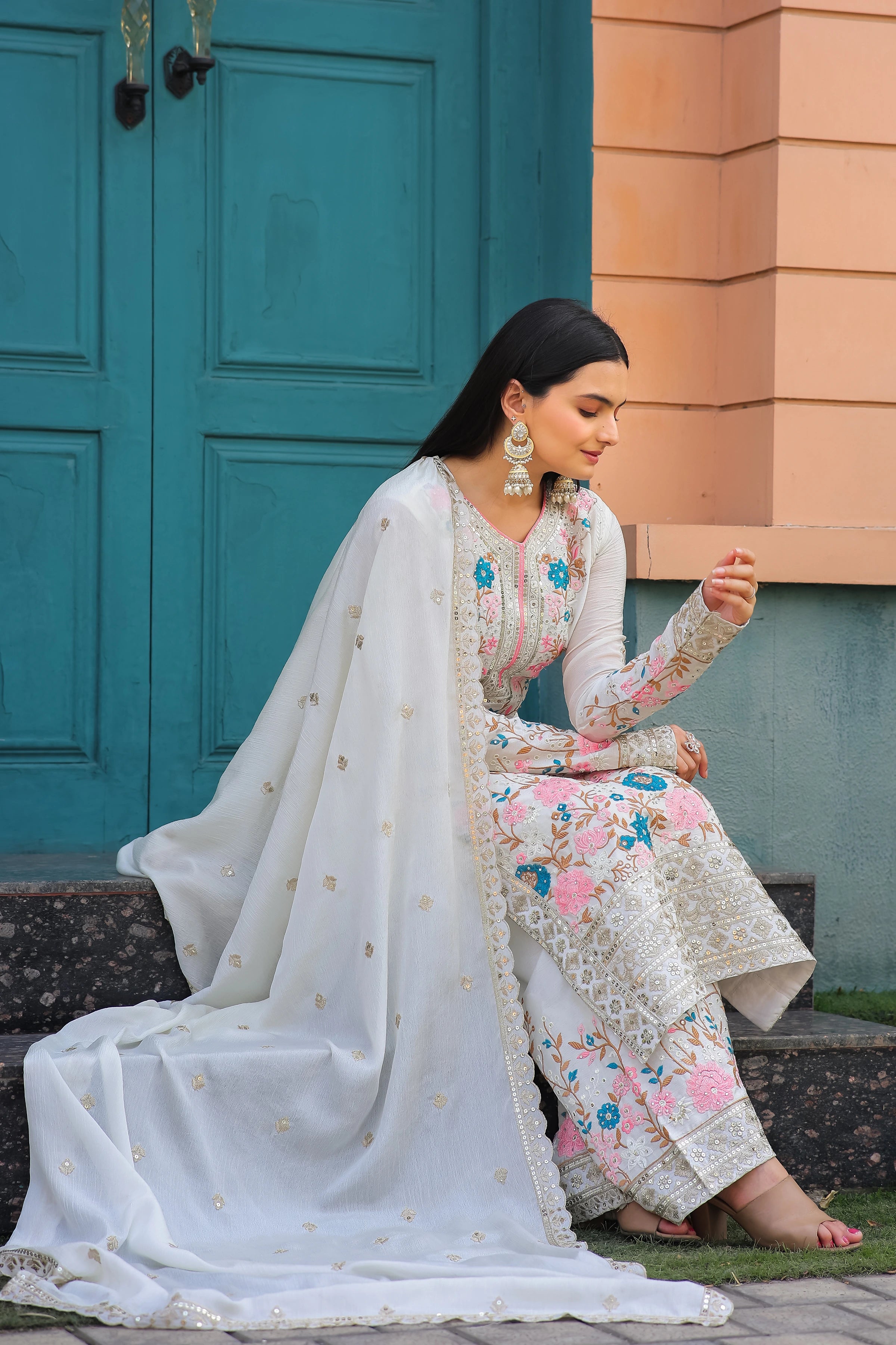 Woman in traditional outfit sitting on steps with a teal door and beige wall in the background