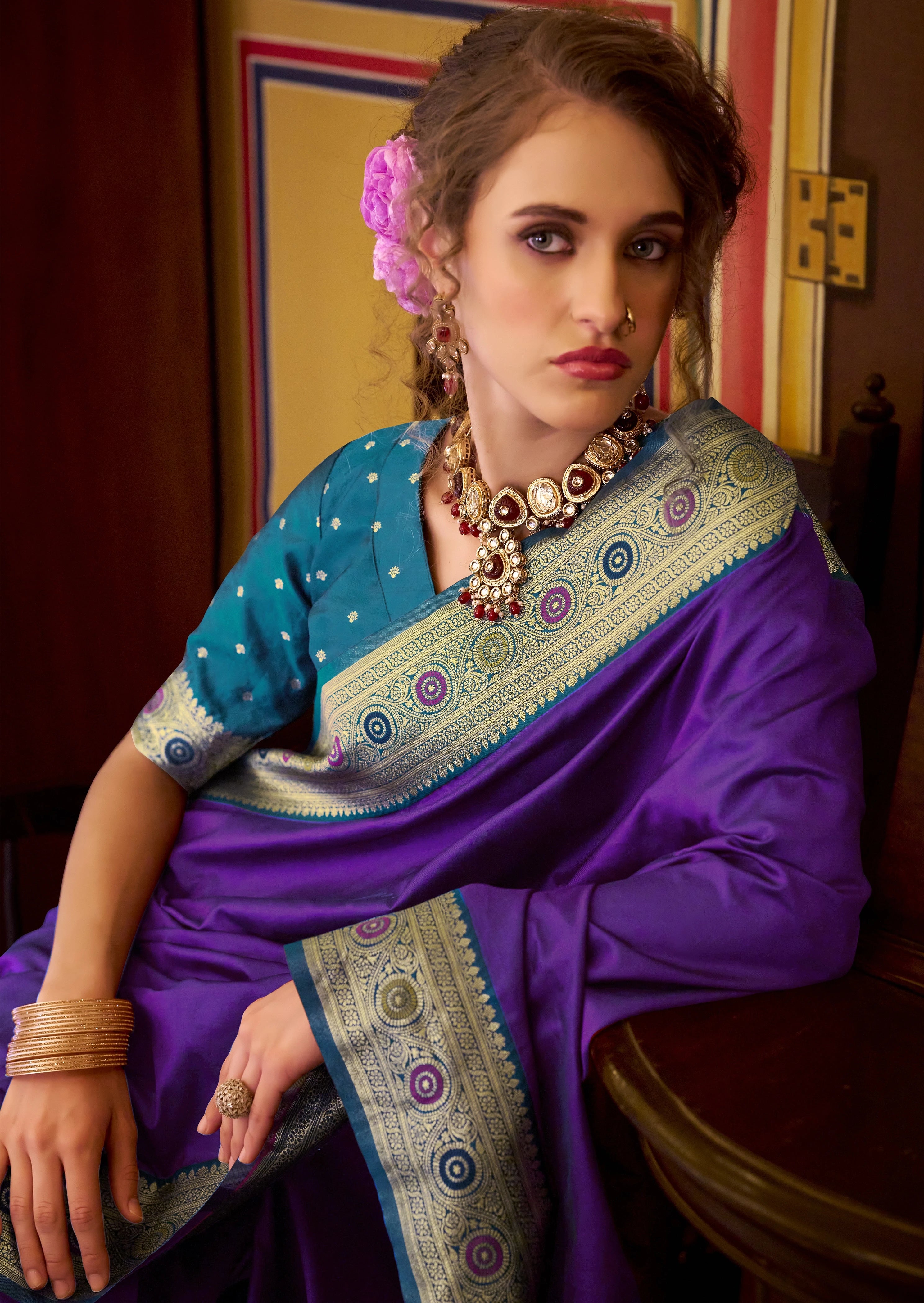 Woman wearing a traditional purple and blue saree with jewelry indoors.