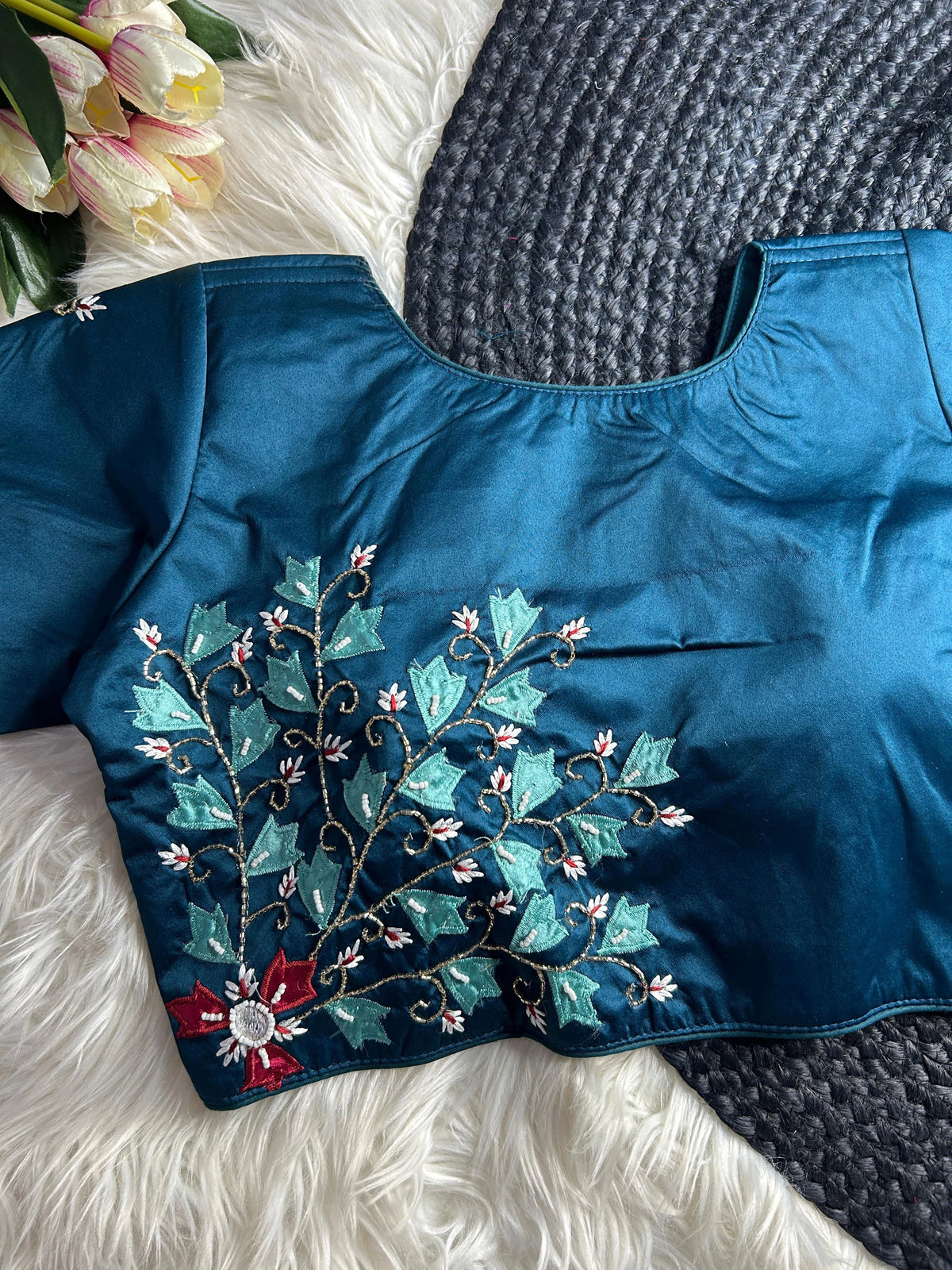 A teal blue silk blouse with a round neckline, featuring intricate hand-embroidery on the lower left side. The embroidery depicts a climbing vine with light blue leaves, gold beaded stems, and small white floral accents, anchored by a larger red flower with a mirrored center at the bottom. The blouse is laid flat on a white faux-fur surface next to a gray woven mat and a bunch of pink and white tulips.