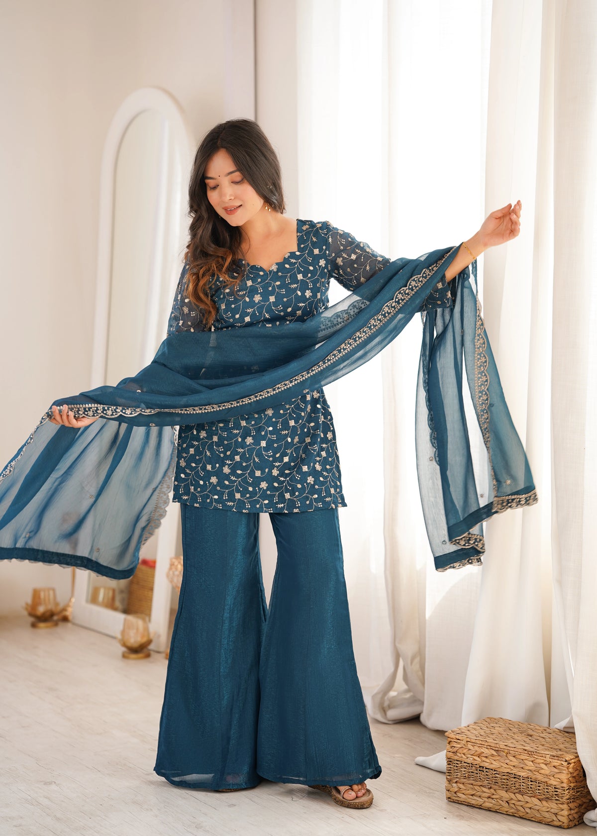 Woman in a blue traditional outfit holding a matching scarf in a bright room.