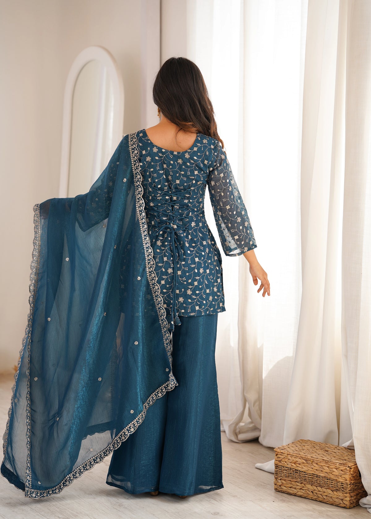 Woman in a teal traditional outfit with a matching dupatta standing in a bright room.