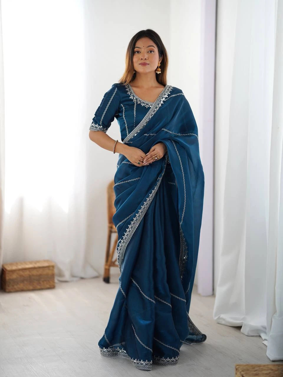 Woman wearing a blue saree with white patterns in a room with white curtains.