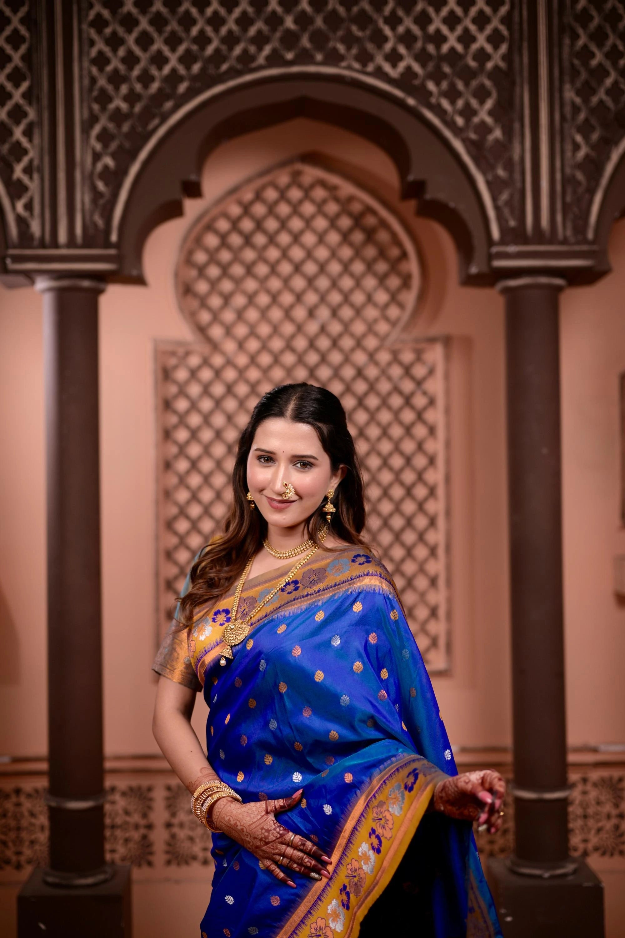 Woman in a blue saree with gold patterns standing in front of decorative arches.