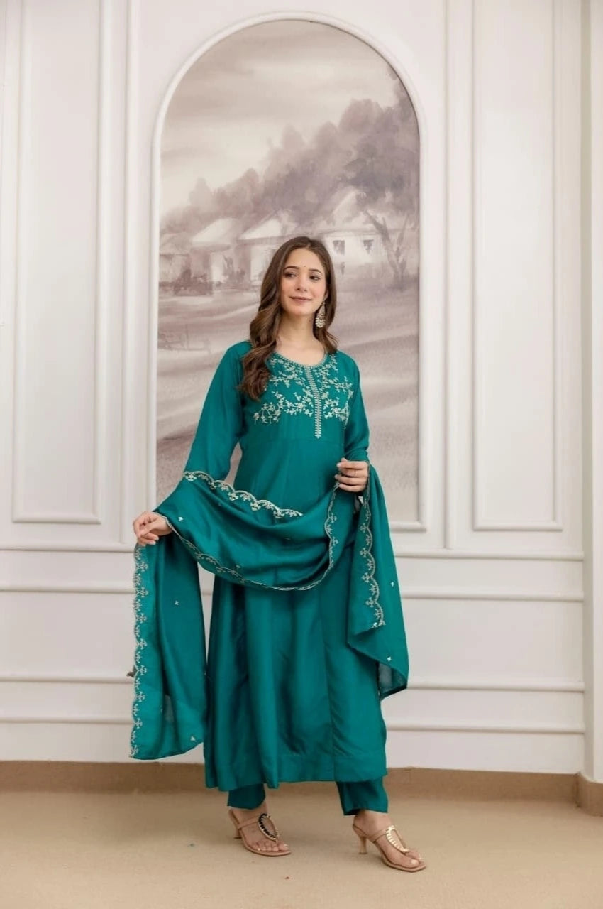 Woman in a teal embroidered outfit standing in front of a decorative wall mirror.