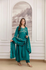 Woman in a teal embroidered outfit standing in front of a decorative wall mirror.