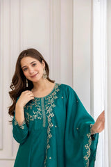 Woman wearing a teal dress with silver embroidery against a white curtain background