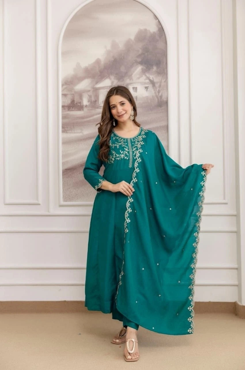 Woman in a teal traditional outfit with silver embroidery standing in a room with white walls and a decorative mirror.