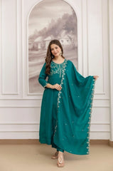 Woman in a teal traditional outfit with silver embroidery standing in a room with white walls and a decorative mirror.