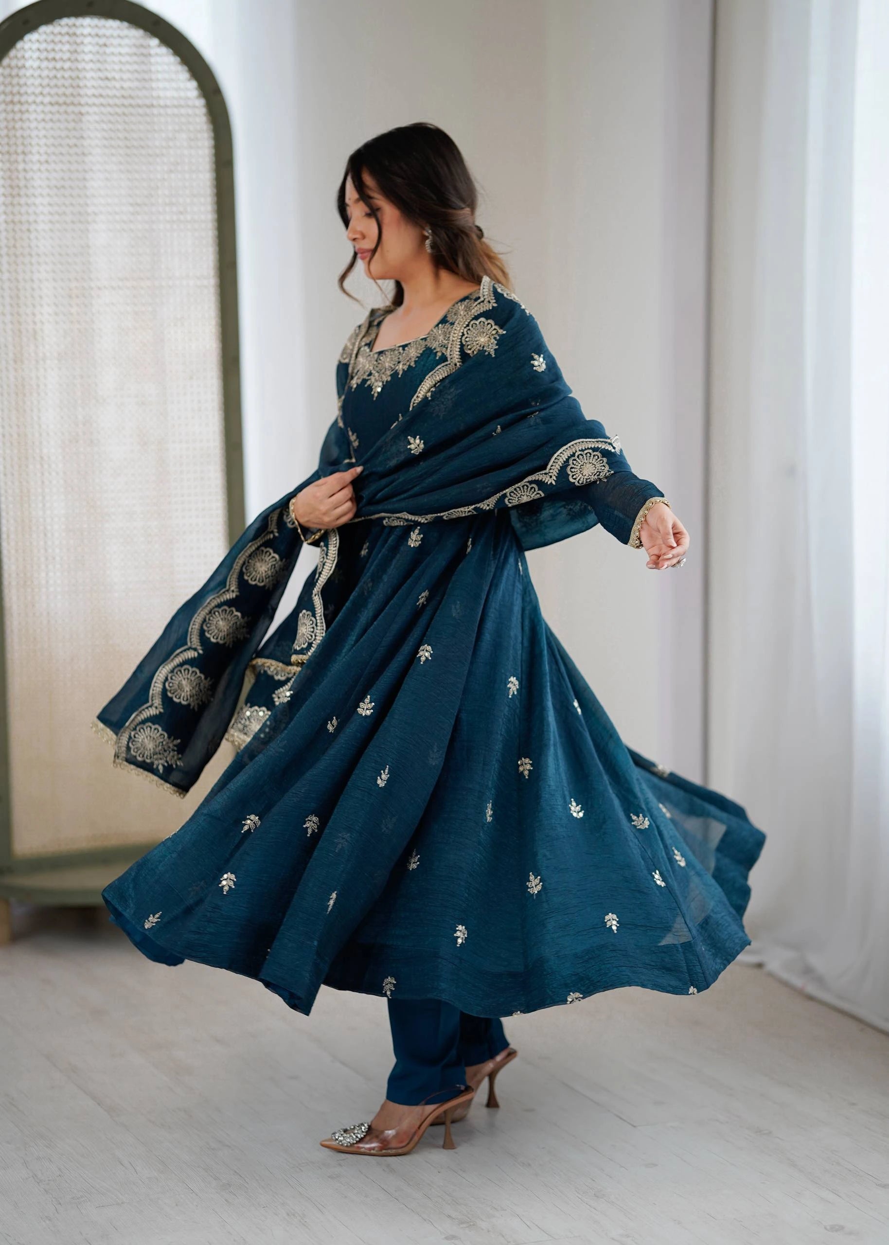 Woman in a teal embroidered outfit standing indoors with a white curtain background