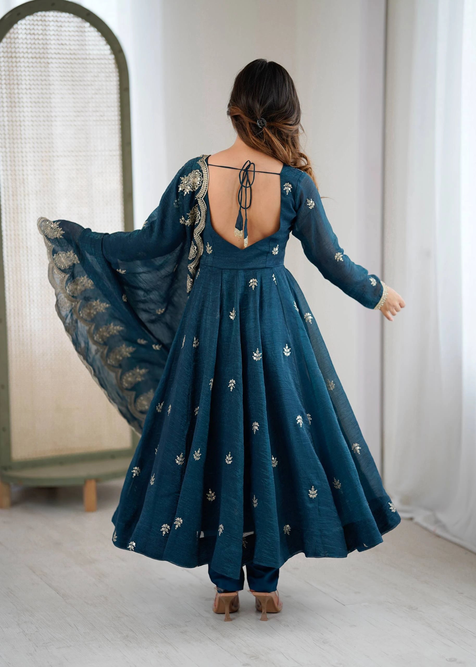 Woman in a teal embroidered dress with a flowing train, standing in a room with white walls and a mirror.