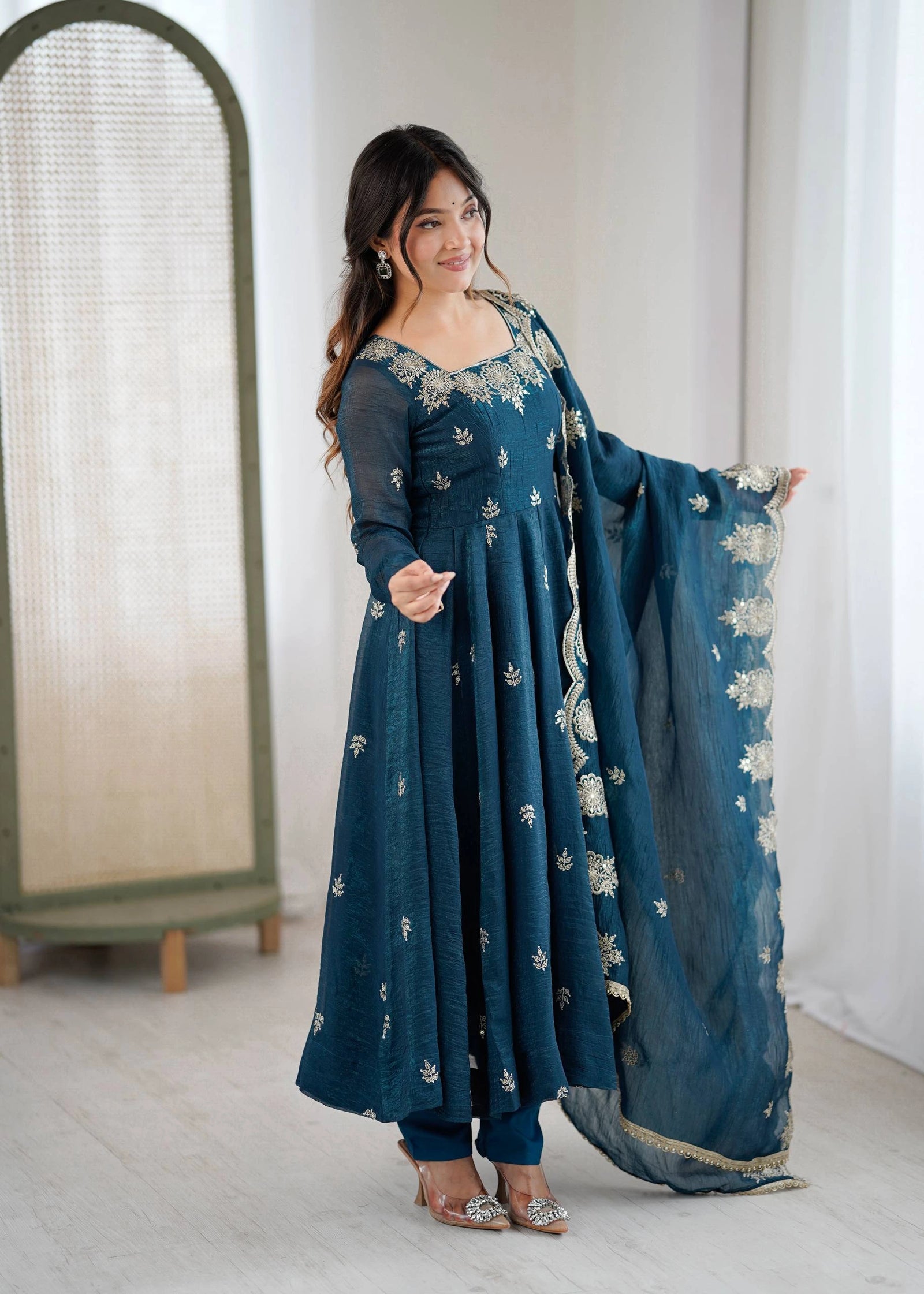 Woman wearing a teal embroidered outfit with a matching dupatta in a minimal indoor setting.