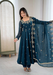 Woman wearing a teal embroidered outfit with a matching dupatta in a minimal indoor setting.