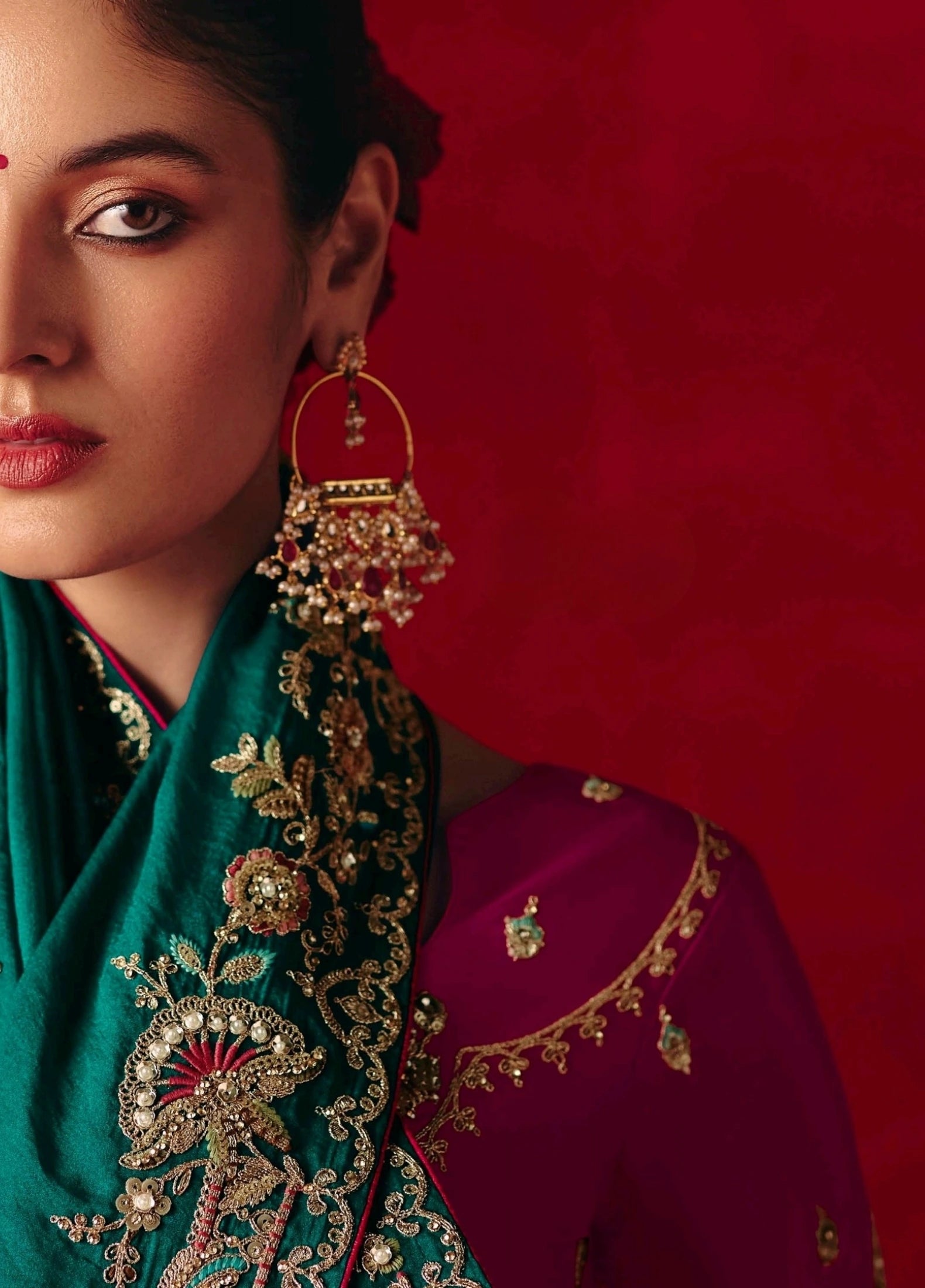 Woman wearing traditional jewelry and a green and red embroidered outfit against a red background