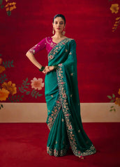 Woman wearing a teal saree with floral patterns against a red and beige background