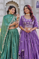 Two women in traditional green and purple sarees standing in front of an architectural background.