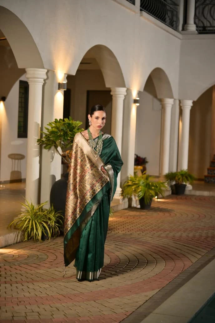 Woman in a green saree standing in an elegant indoor setting with arches and plants.