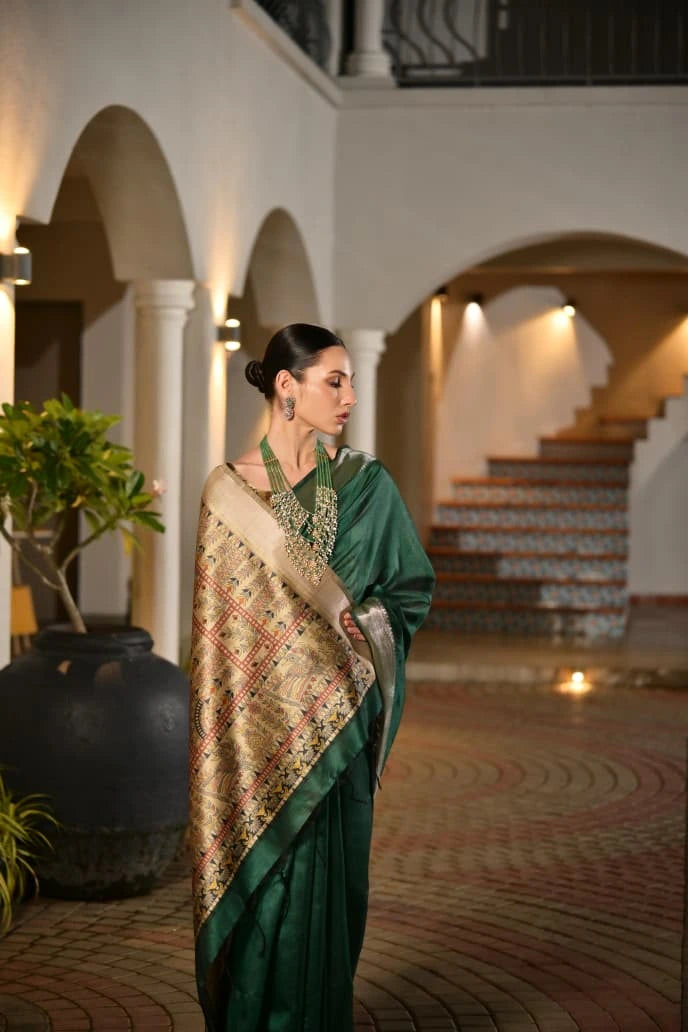 Woman in a green saree with a patterned shawl standing in an elegant indoor setting.