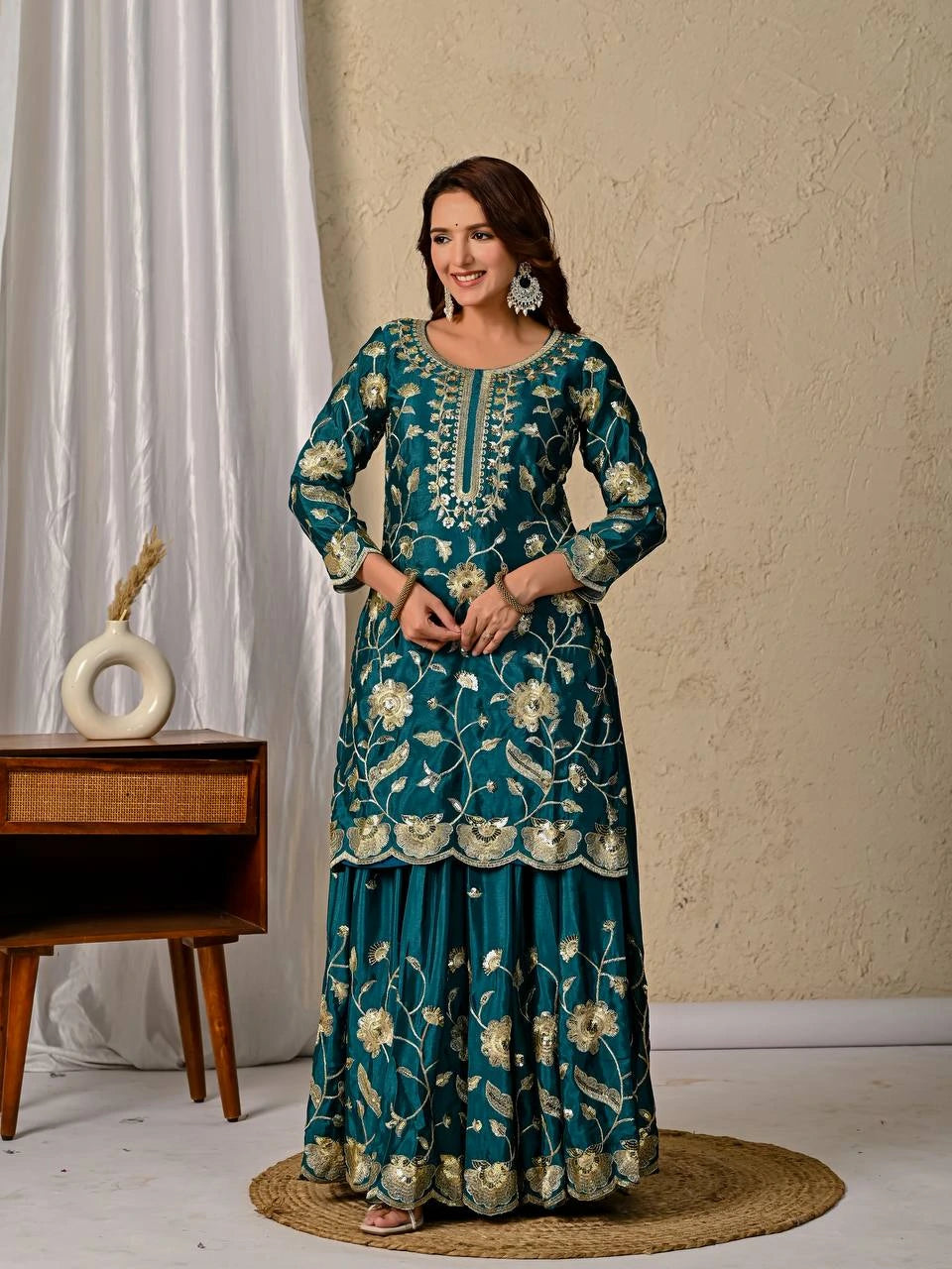 Woman wearing a teal embroidered traditional outfit standing in a room with a neutral background.