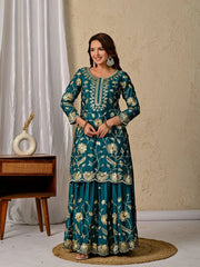 Woman wearing a teal embroidered traditional outfit standing in a room with a neutral background.