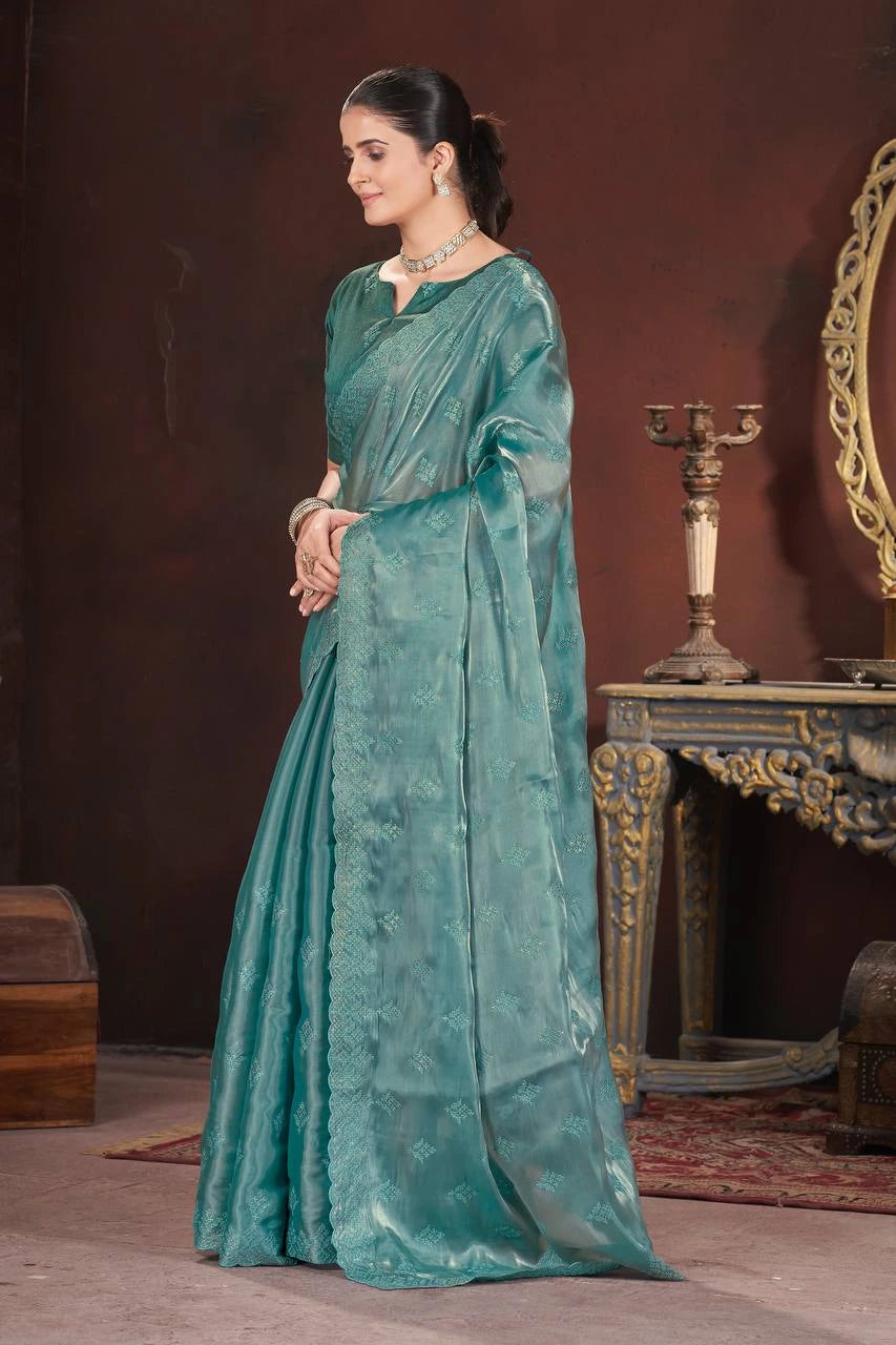 Woman in a teal saree standing in a room with a dark brown wall and decorative elements.