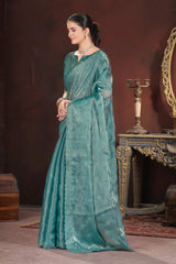 Woman in a teal saree standing in a room with a dark brown wall and decorative elements.