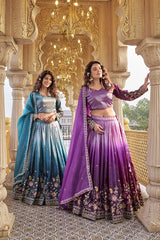Two women in traditional blue and purple lehengas standing in an ornate architectural setting.