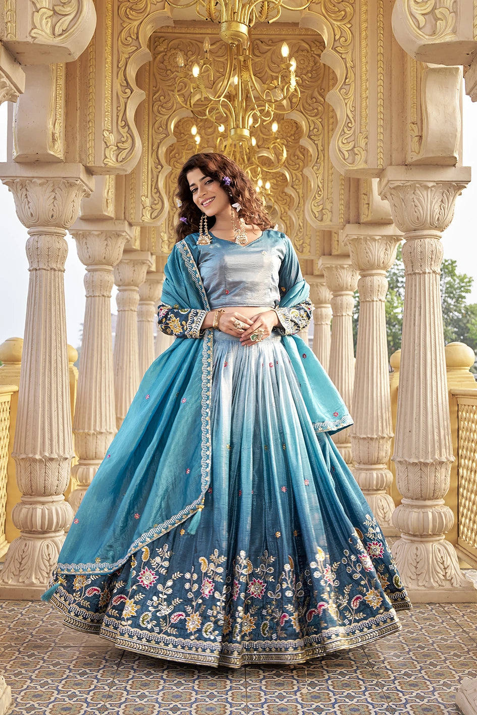 Woman in a blue embroidered traditional outfit standing in an ornate architectural setting.