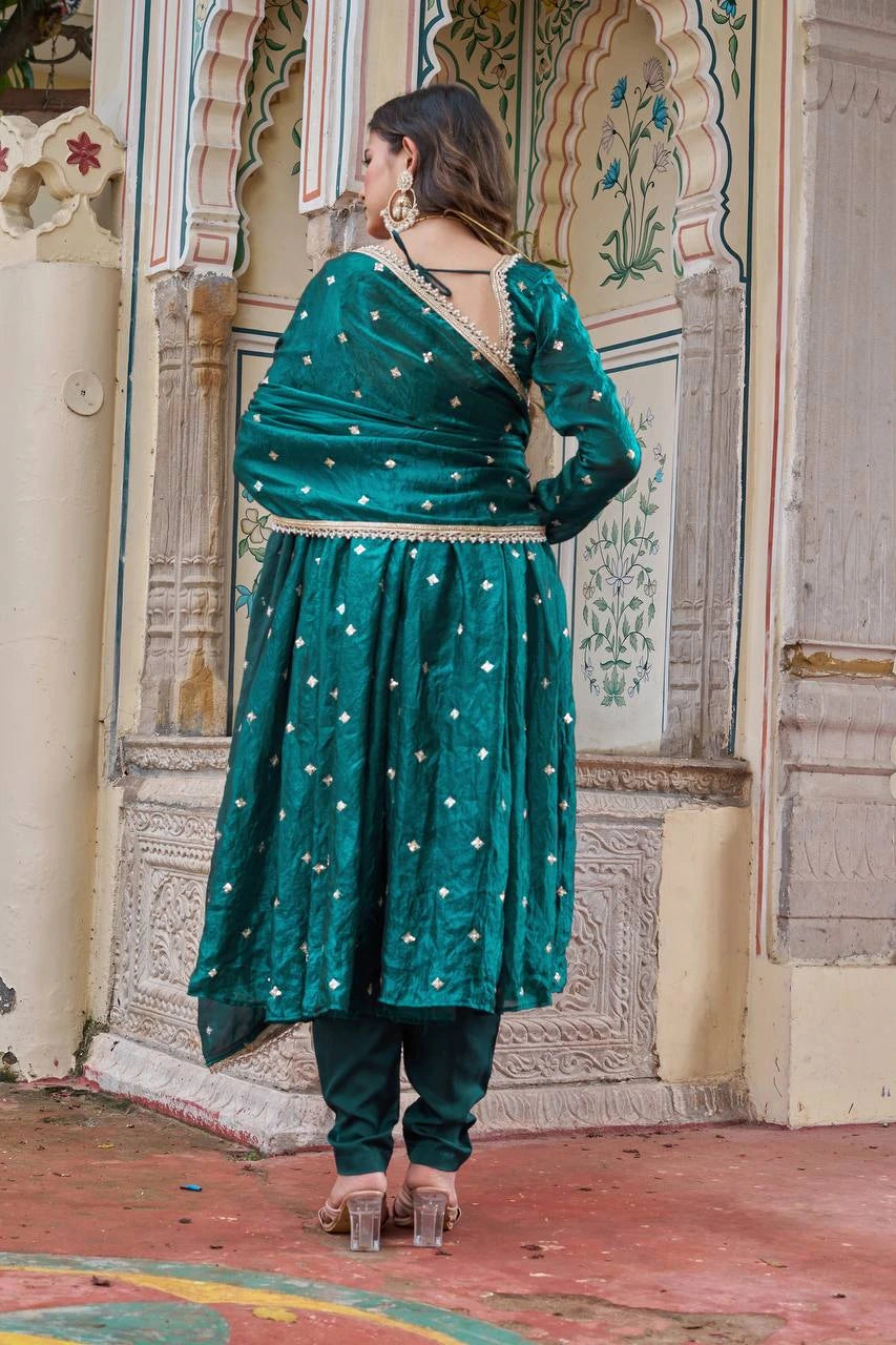 Woman in a teal traditional outfit standing in front of decorative wall.