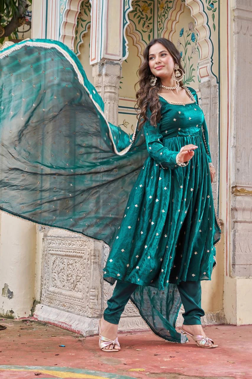 Woman in a teal traditional outfit with a flowing dupatta in front of an ornate architectural background.