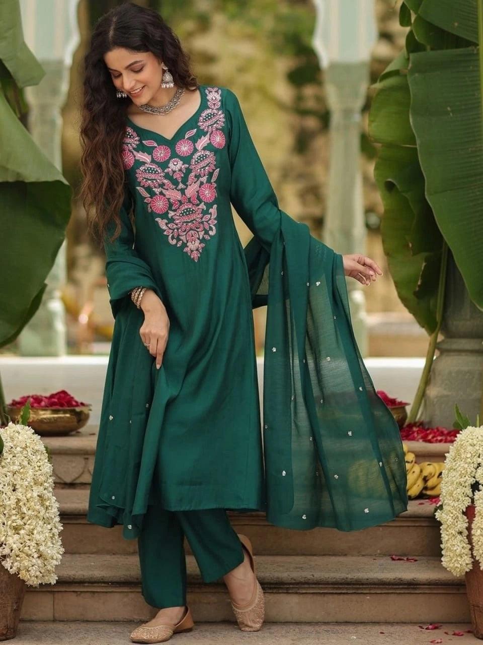 A detailed close-up of a dark green fabric, likely the sleeve of a kurti, showcasing a fine texture and scattered small, silver floral-shaped sequin embellishments. In the background, a portion of vibrant pink and gold floral embroidery on the main garment is softly blurred.