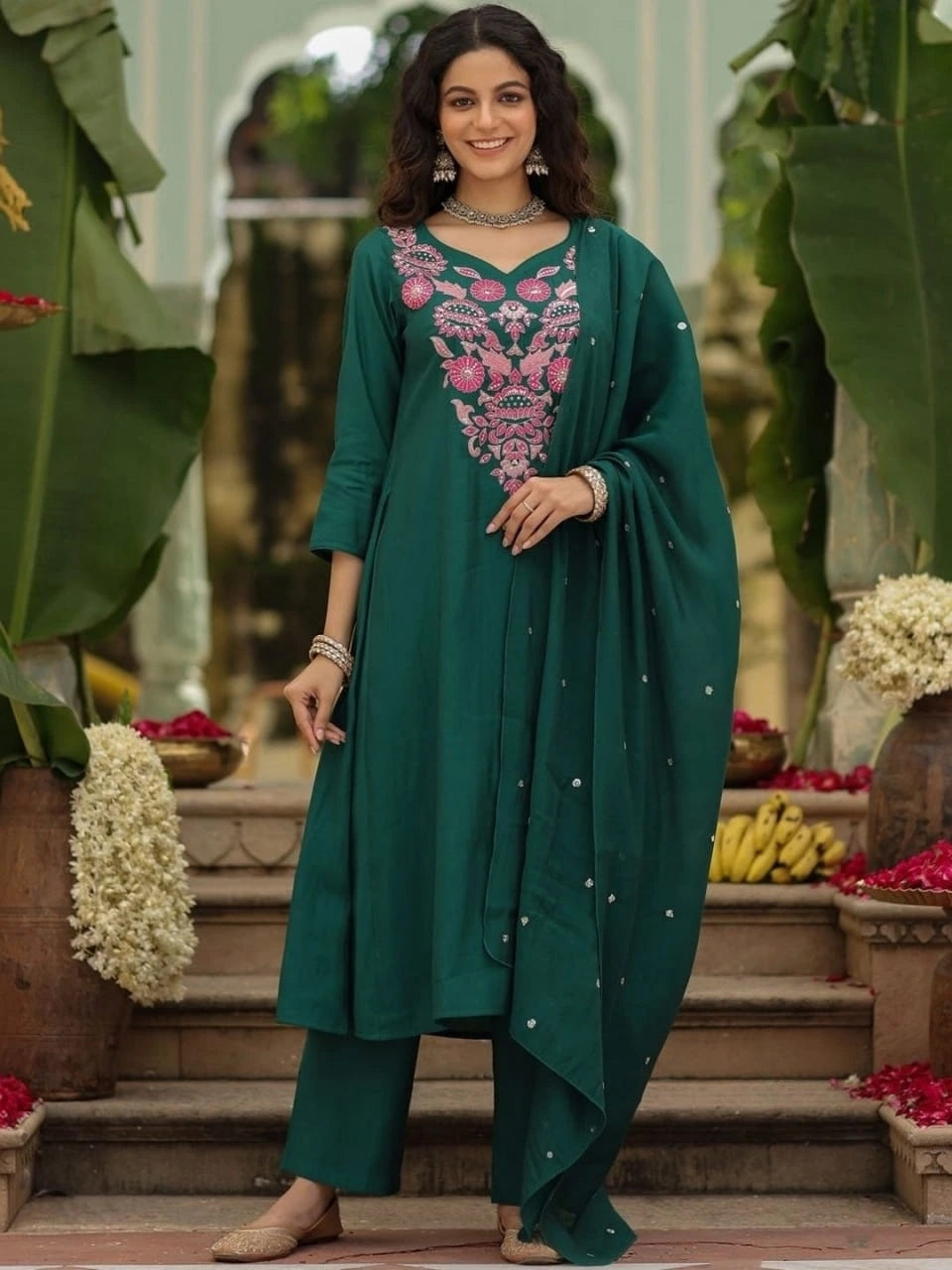 Woman in a teal traditional outfit with pink embroidery standing outdoors.