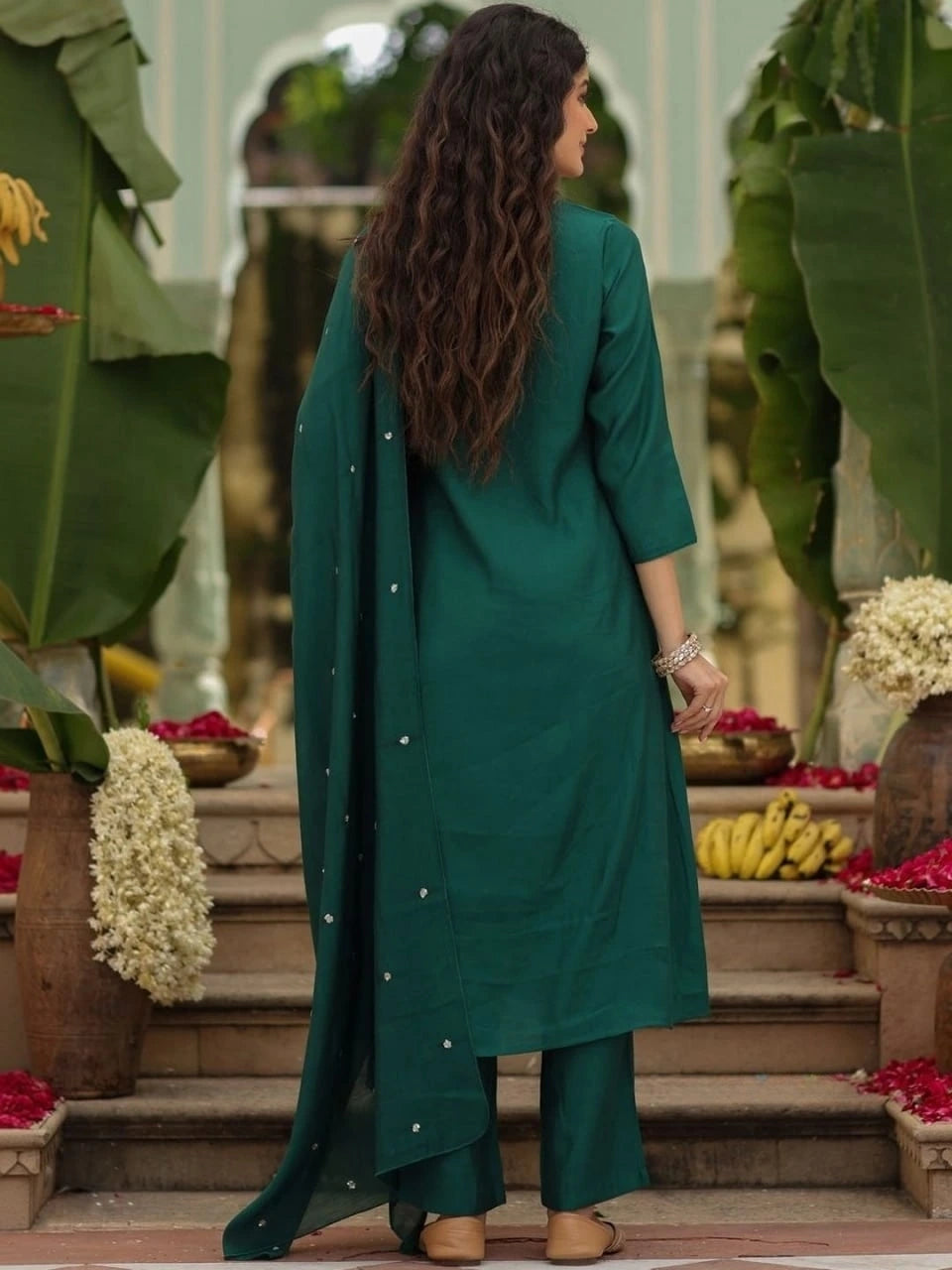 Woman in a green outfit standing in an outdoor setting with plants and flowers.