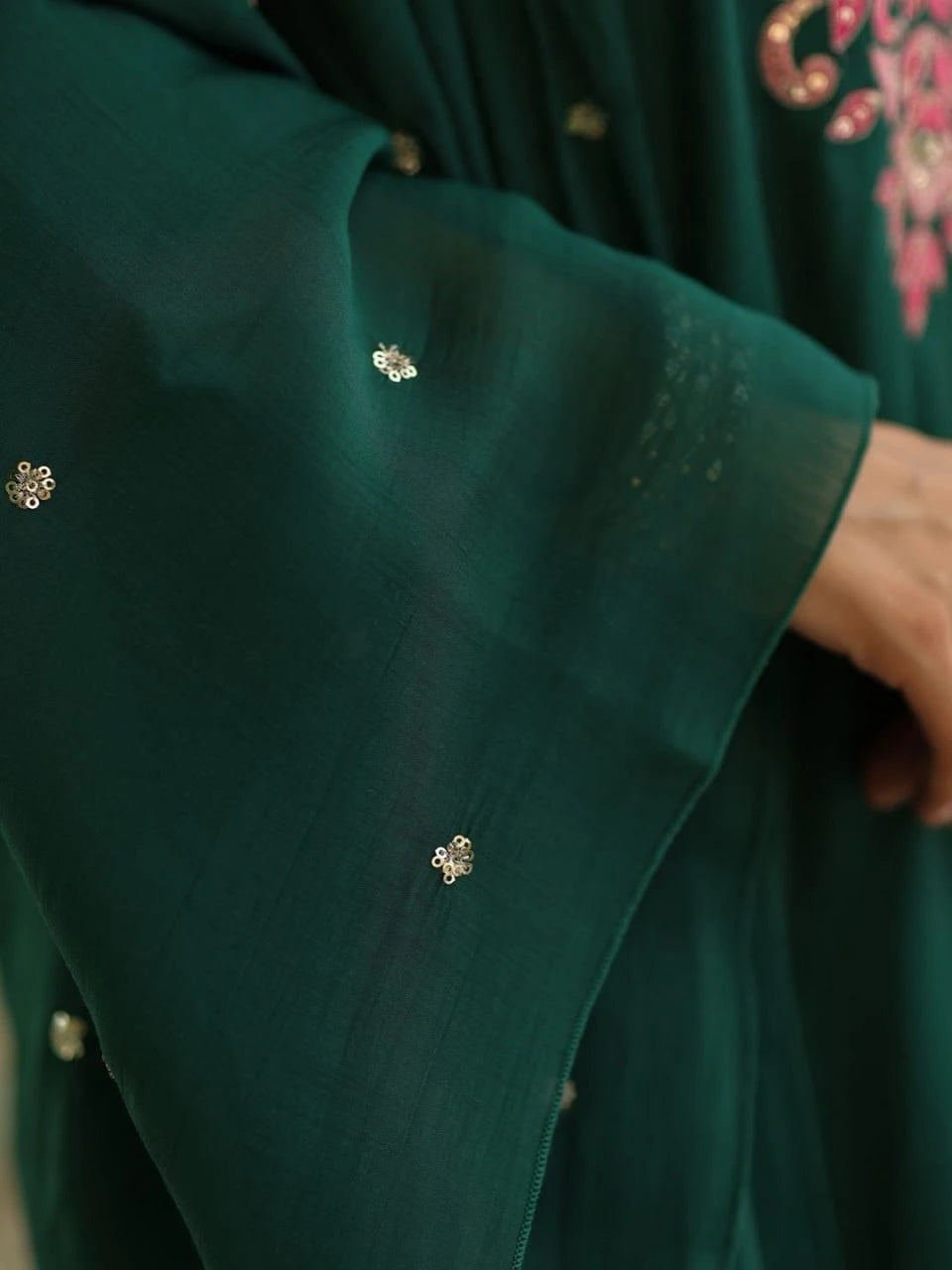 A woman stands on stone steps wearing a dark green ethnic suit. The long kurti features an intricate V-neckline with pink and light purple floral embroidery. She is styled with matching straight-cut trousers, a sheer dupatta with tiny sequins, silver oxidized jewelry, and beige juttis. The scene is decorated with large green leaves and white floral garlands.