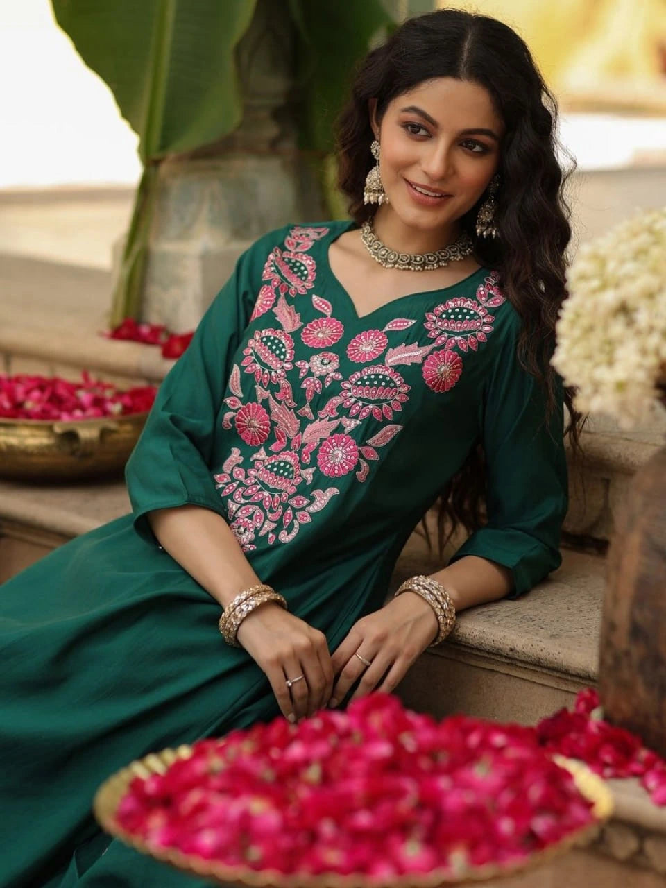 Woman in a green embroidered dress sitting on a step with flowers around her.