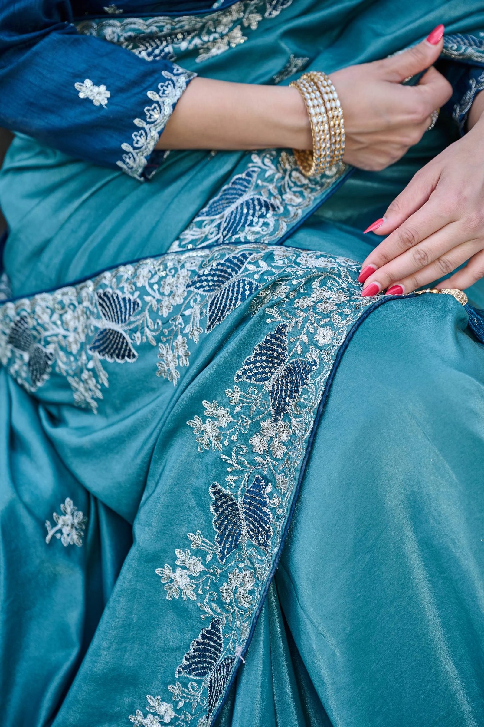 Close-up of a teal embroidered garment with gold jewelry and red nail polish.