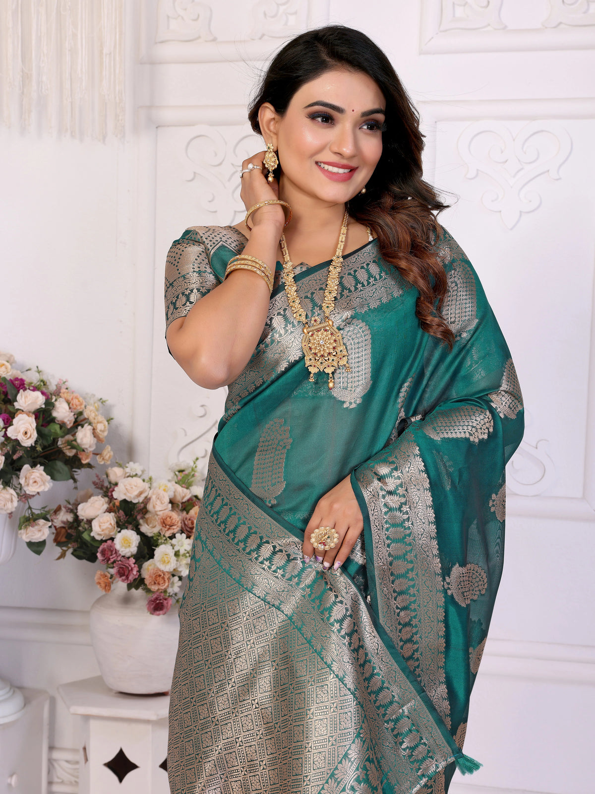 Woman wearing a green and gray saree with jewelry, standing against a white wall with floral decorations.
