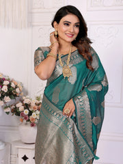 Woman wearing a green and gray saree with jewelry, standing against a white wall with floral decorations.