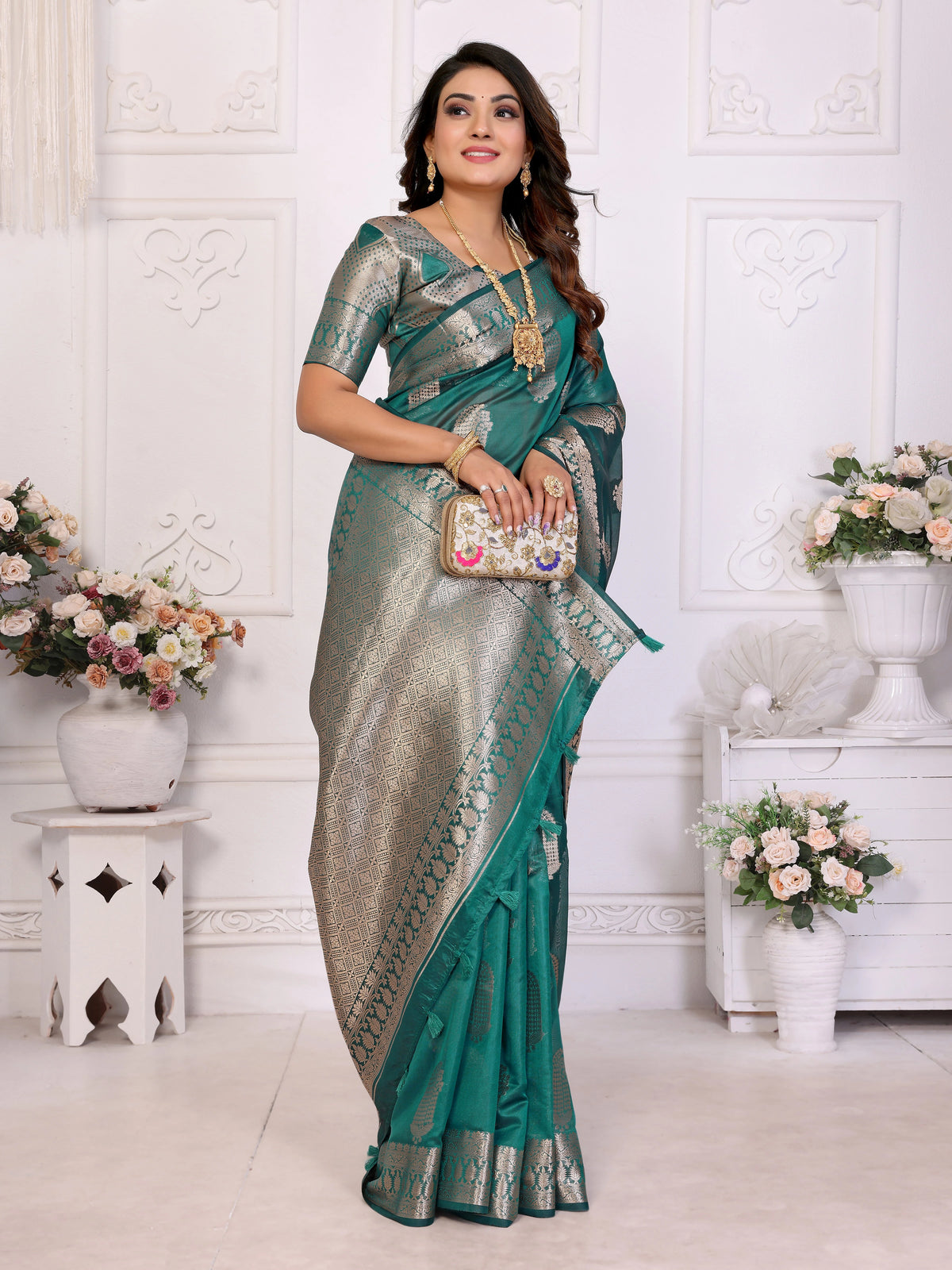 Woman wearing a green and silver saree in a decorated room with flowers and white walls.