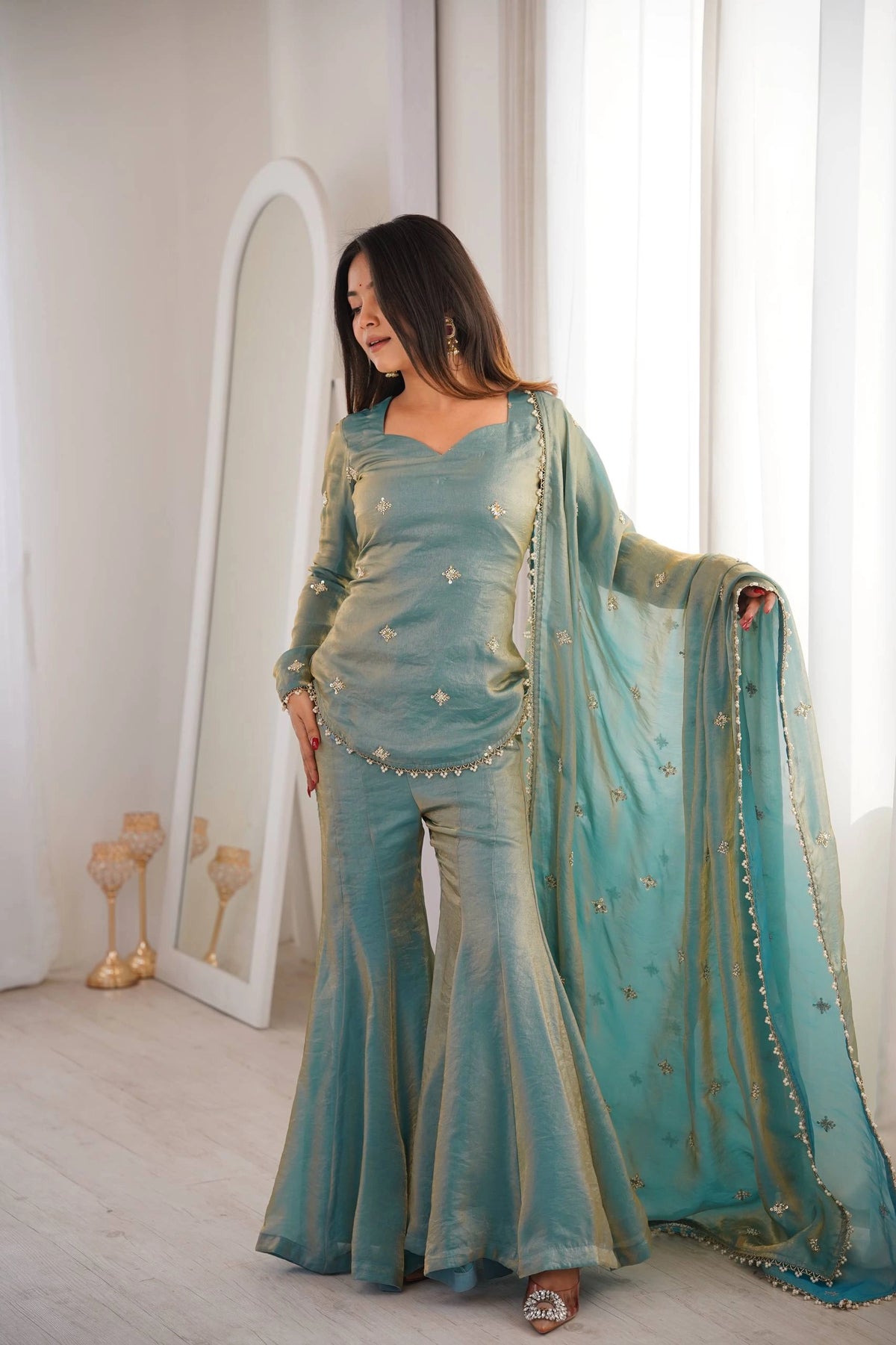 Woman in a teal traditional outfit standing in a well-lit room with a mirror and decorative items.