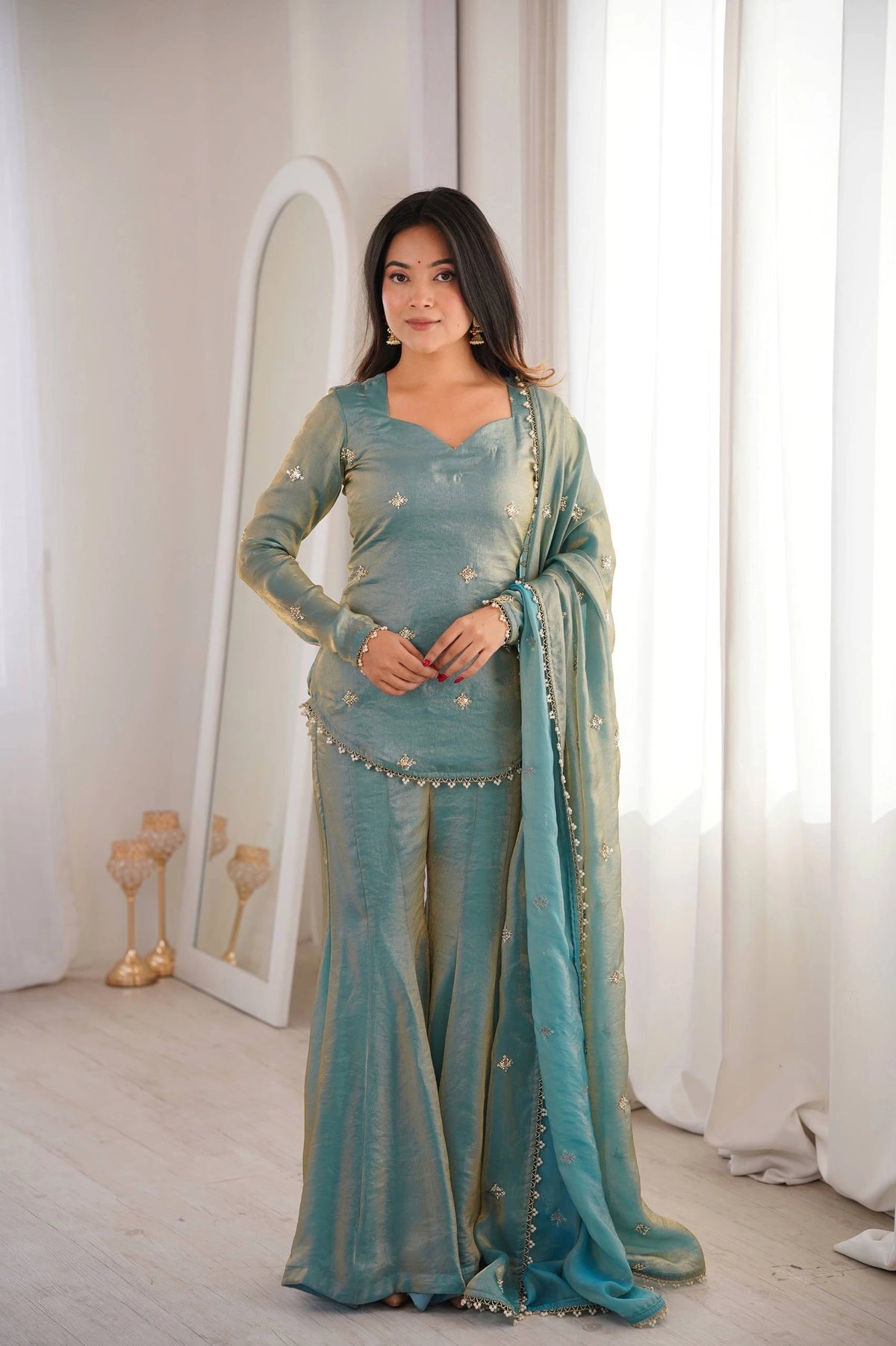 Woman in a teal traditional outfit standing in a room with white curtains and decor.