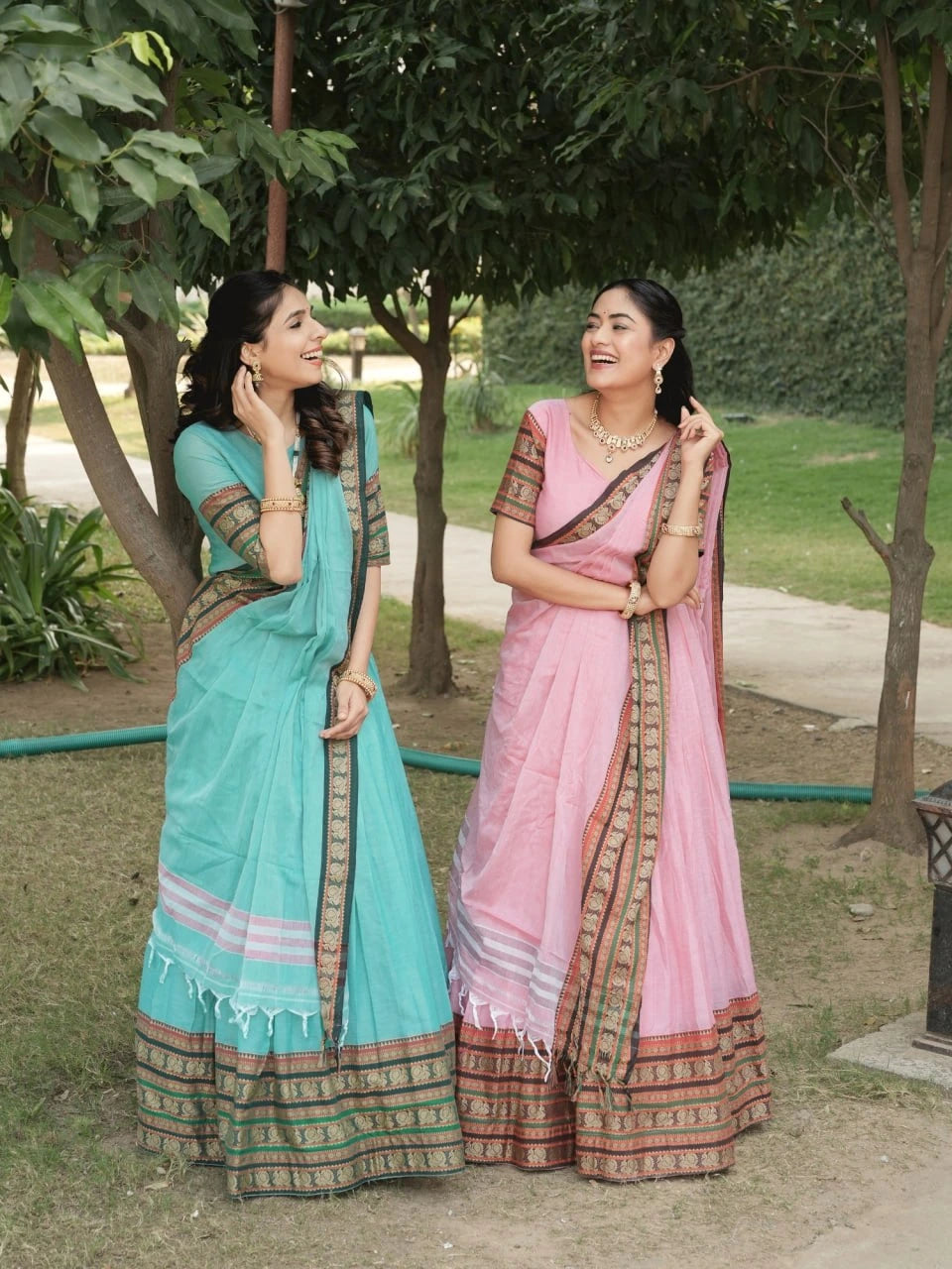 Two women in traditional sarees standing outdoors with trees and grass in the background.