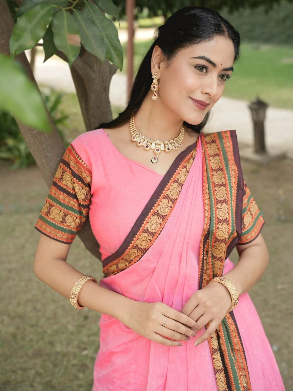 Woman wearing a pink saree with gold and green patterns outdoors.