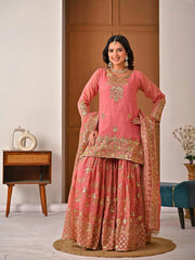 Woman in a traditional pink and gold embroidered outfit standing in a room with wooden furniture.