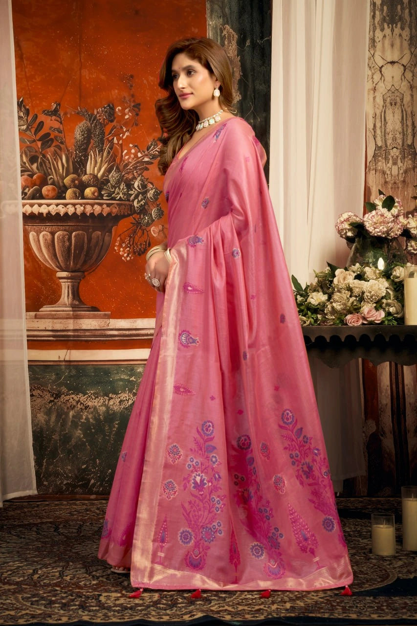 Woman in a pink saree with floral patterns standing in an elegant room.