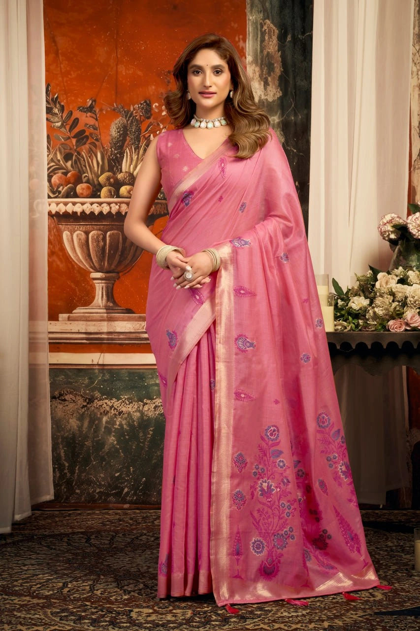 Woman in a pink saree with floral patterns standing in an elegant indoor setting.