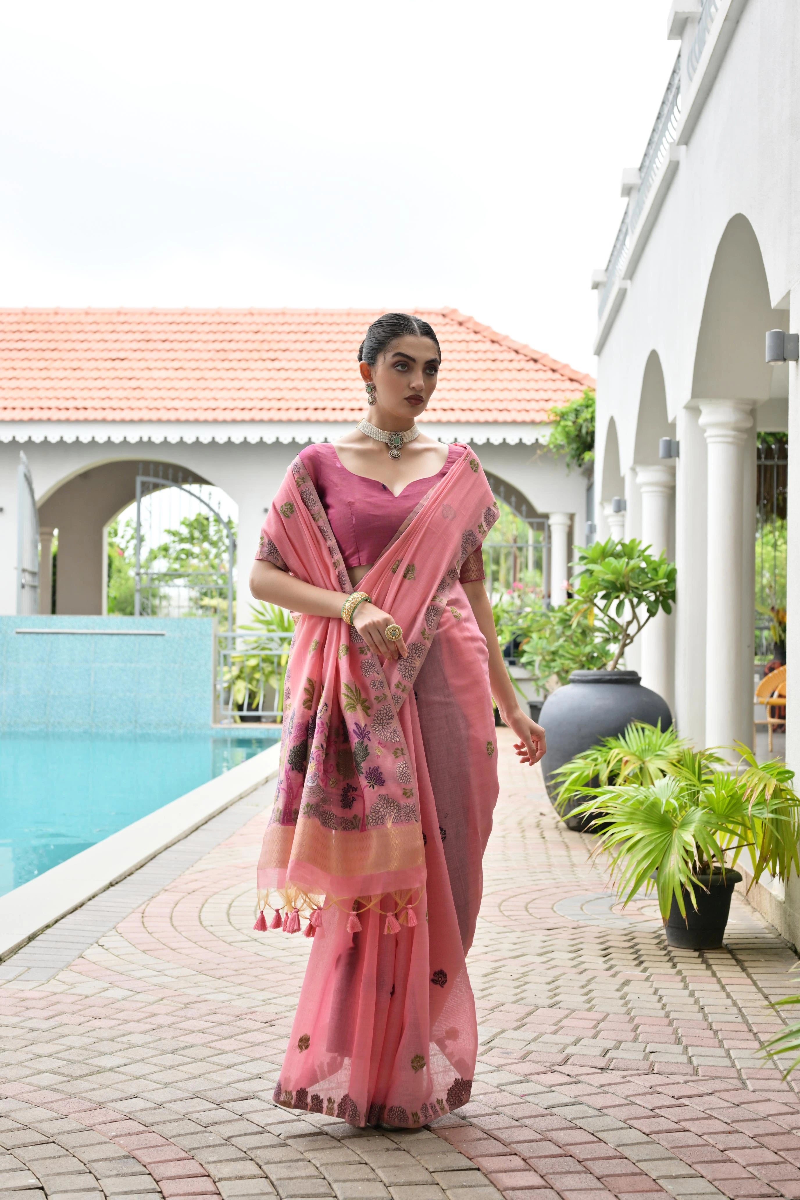 Woman in a pink saree standing by a poolside with a white building and plants in the background.